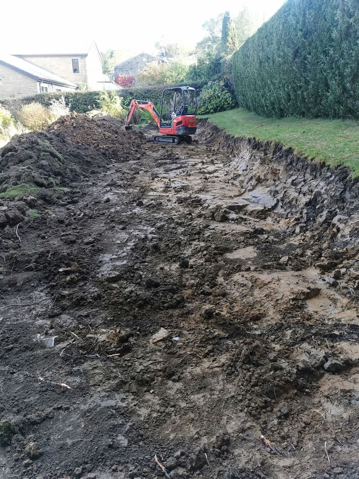 A mini digger working across the stripped slope with spoil banked up behind it
