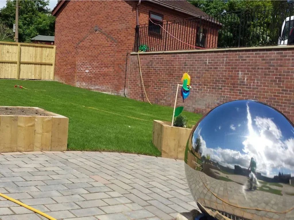 The block-paved path beside timber sleeper planters, with a mirrored sphere on the paving