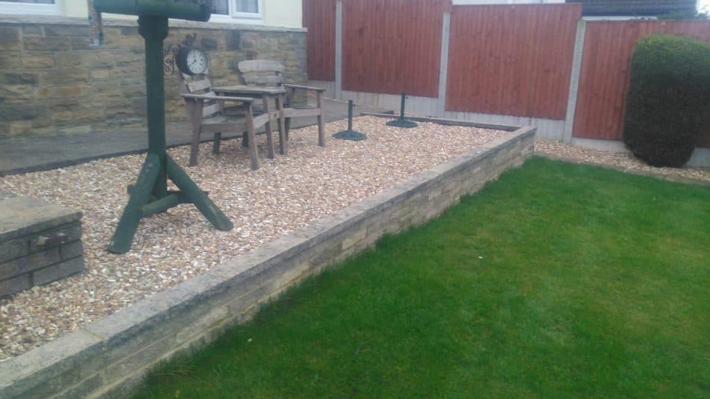 A wooden garden table and chairs set on a gravel bed edged in stone, beside a lawn and a timber fence