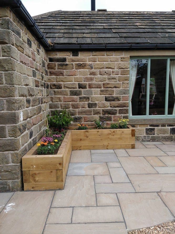 A sleeper-built planter of bedding flowers against a stone house wall, with a sandstone patio laid up to it
