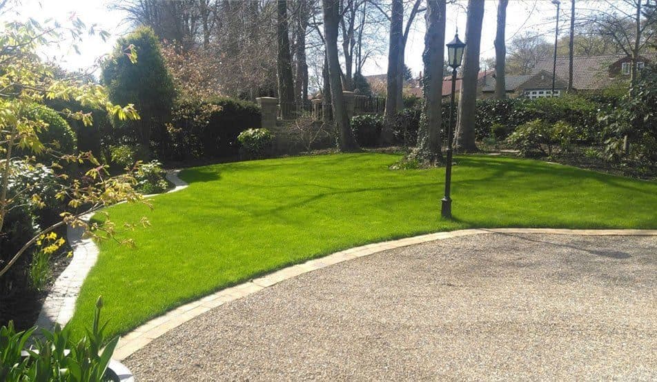 A curving lawn edged with setts beside a gravel drive, with a lamp post and mature trees behind