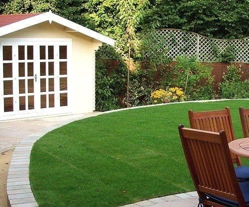 A summer house at the top of a garden with a curved lawn, paved edging path and wooden garden furniture