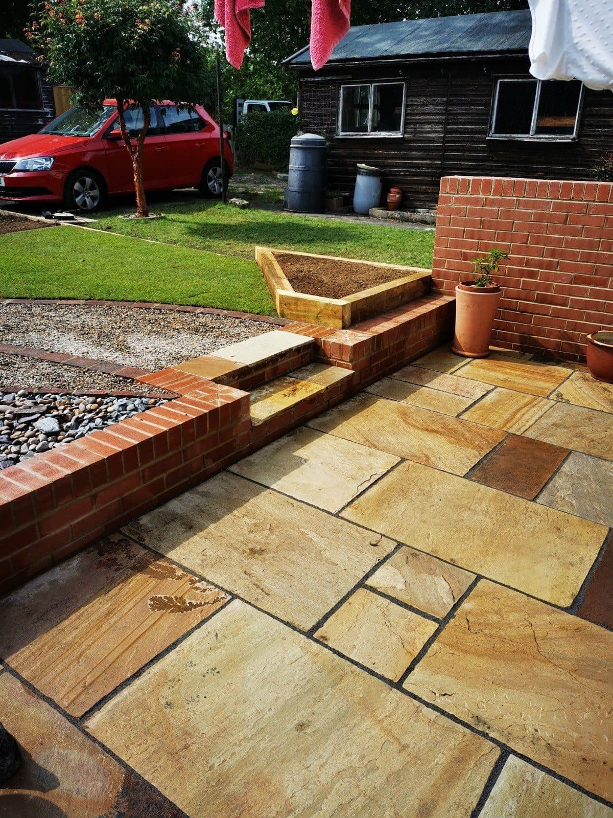A sandstone patio edged with a low brick retaining wall, timber sleeper steps and a sleeper raised bed