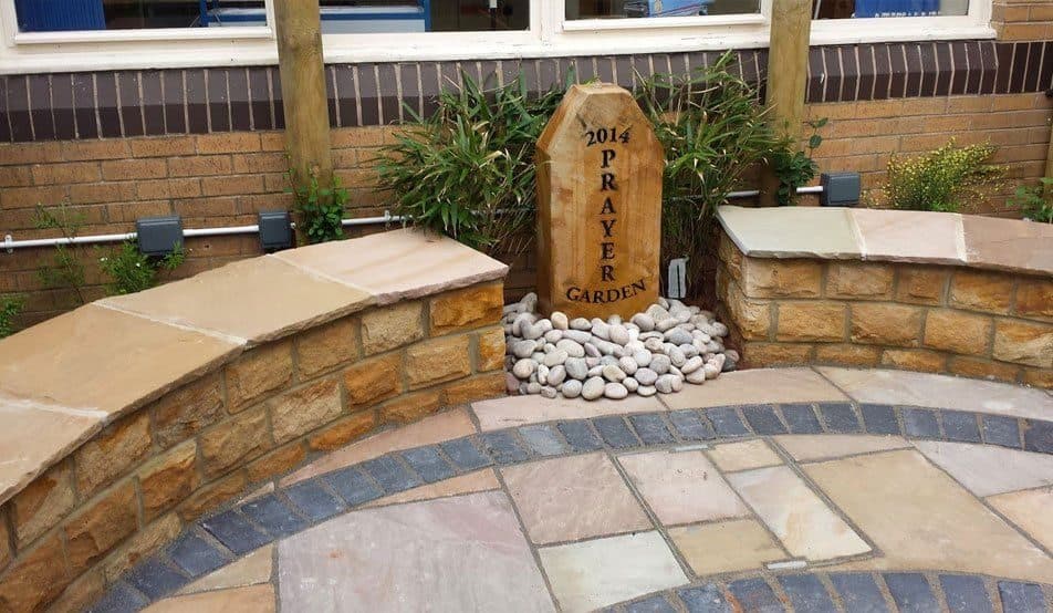 A curved sandstone seat wall around a paved circle of mixed setts and slabs, with a carved stone marker in a pebble bed