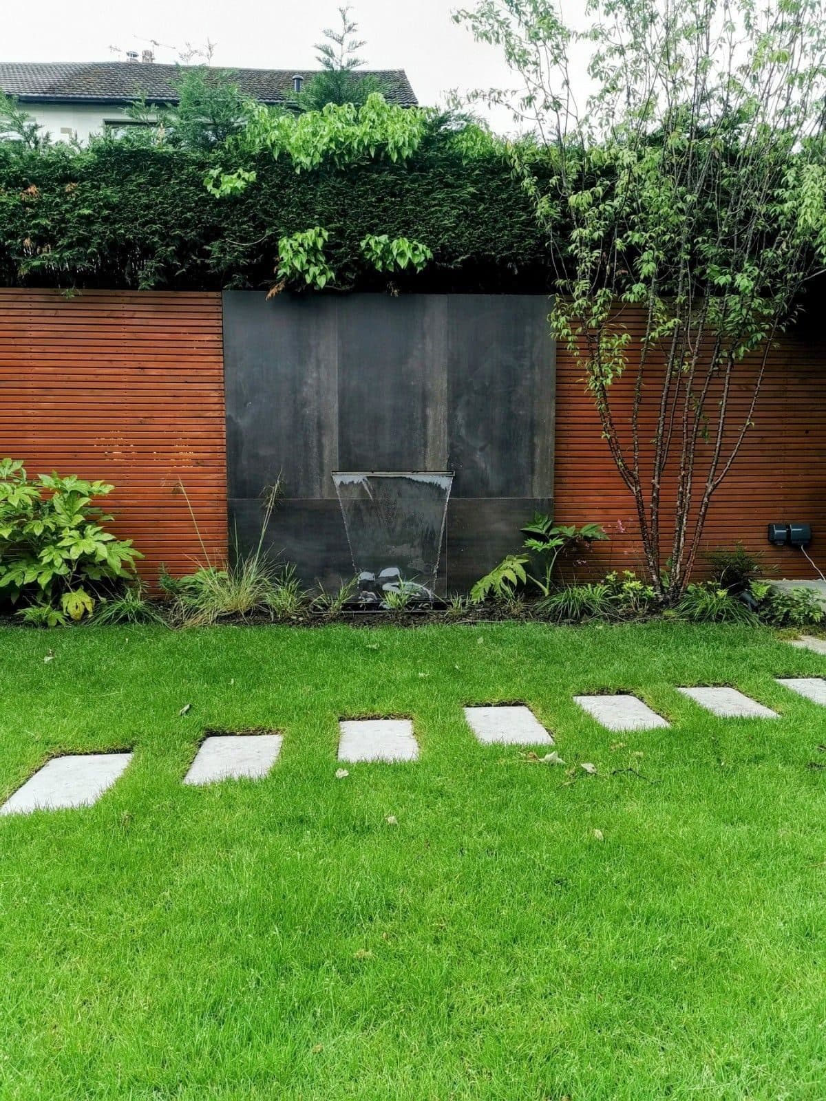 A dark steel water blade set into a slatted timber wall, with paving slabs set as stepping stones across the lawn
