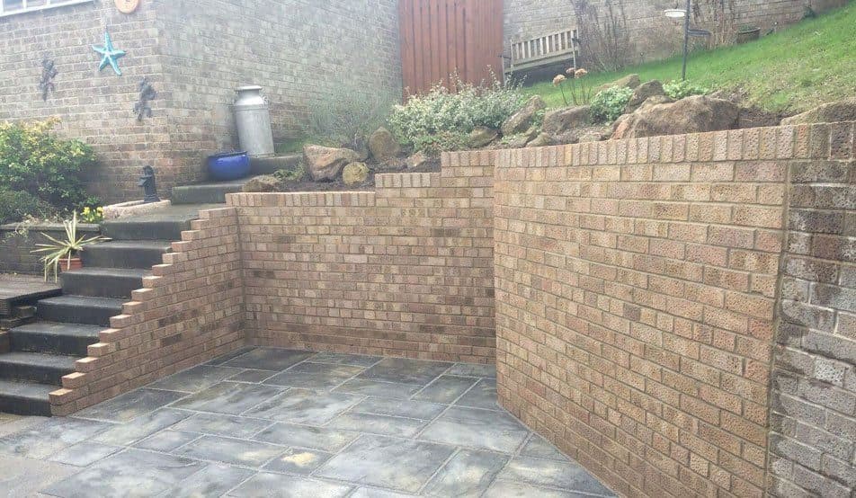 Brick retaining walls holding back a bank around a lower paved terrace, with brick steps running up to the level above