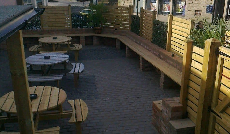 A long timber bench running along slatted timber screens beside a block-paved courtyard set out with wooden tables