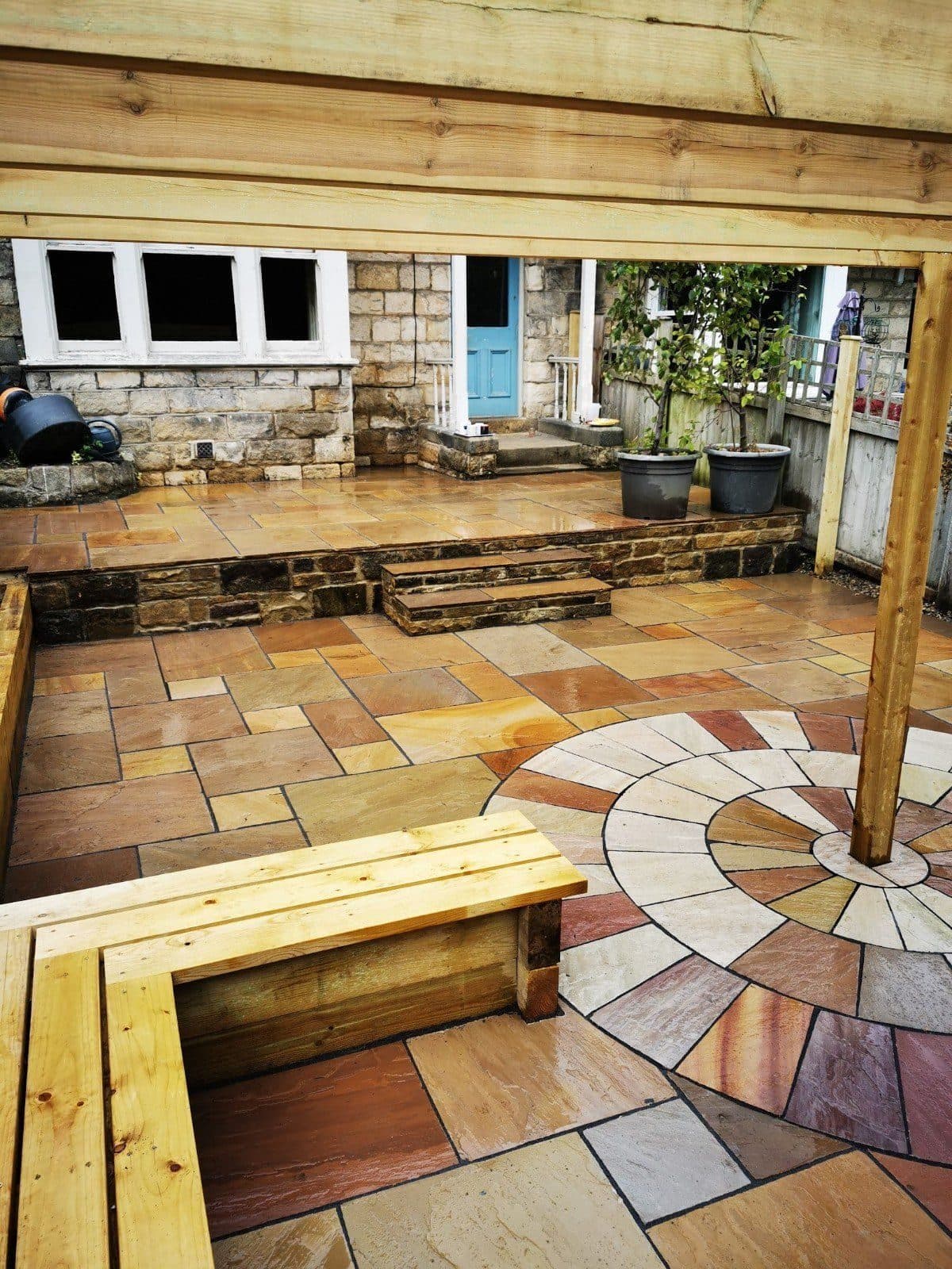 A timber pergola roof over a sandstone patio with a paving circle, a built-in timber bench and steps up to the house