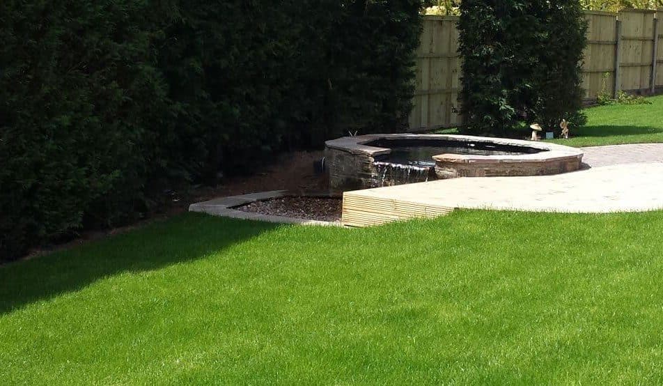 A raised stone pool spilling over a lip into a lower pool, set into a lawn beside a paved terrace and a tall hedge