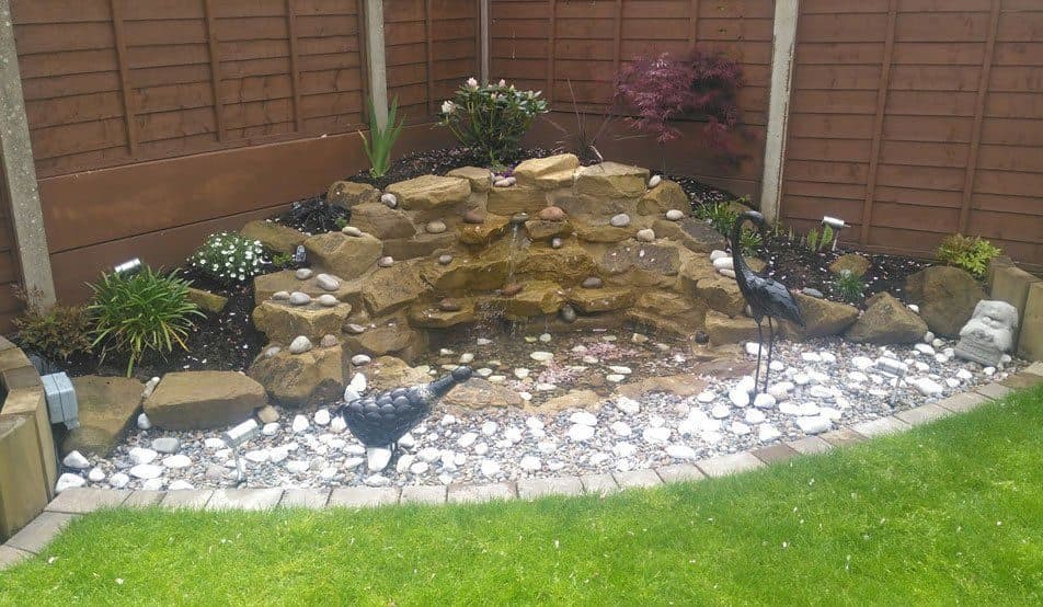 A cascade built from stacked stone in a corner bed, running down into a pebble bed edged with setts