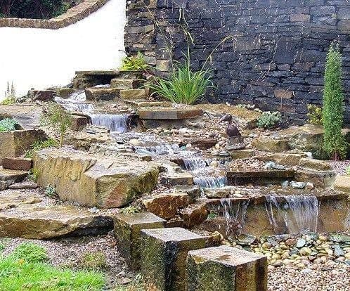A run of stone slabs stepping a waterfall down a steep bank between planted pockets and a stone wall