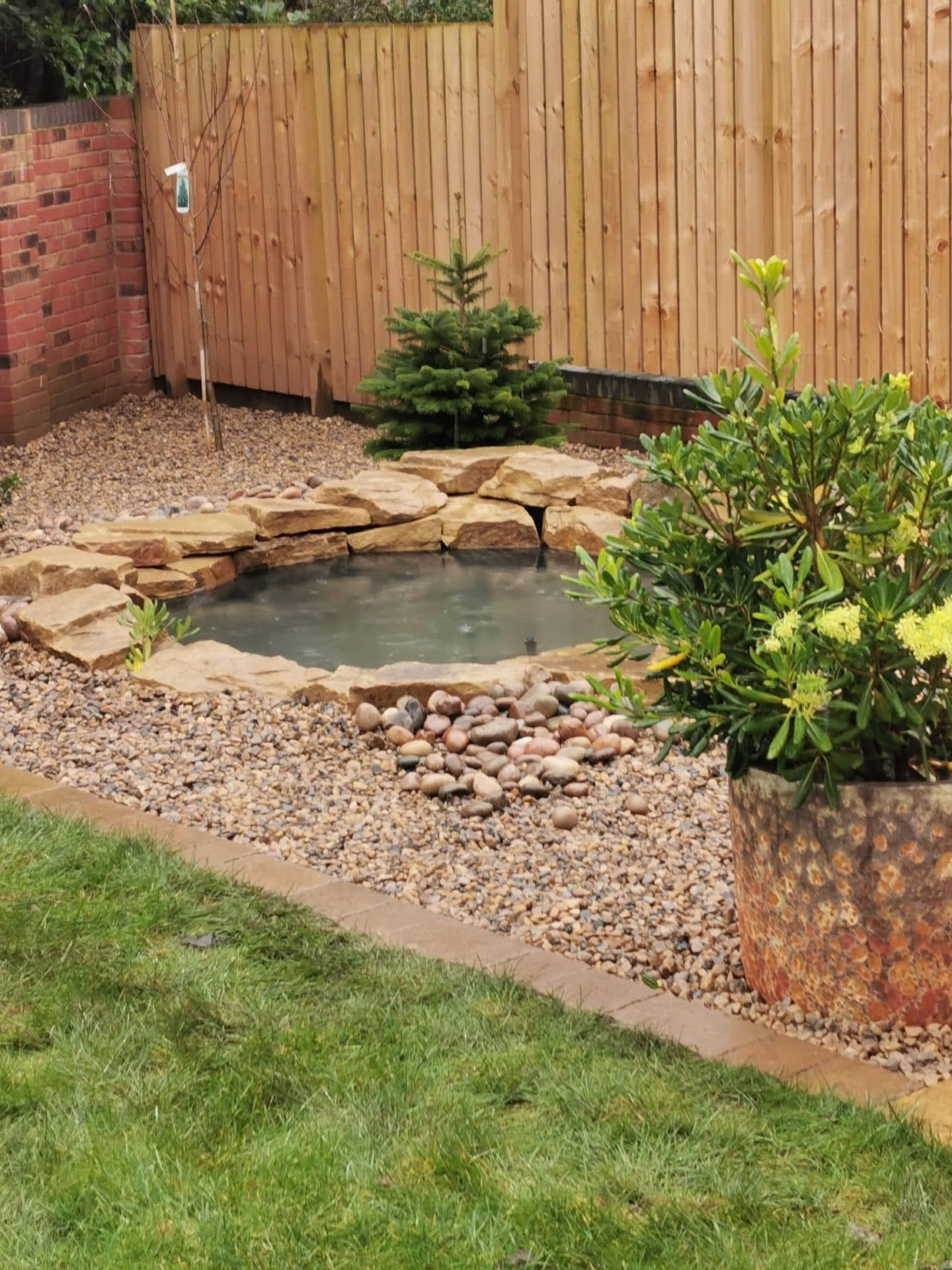 A small pond edged with stone slabs and pebbles, set in a gravel bed against a close-boarded fence